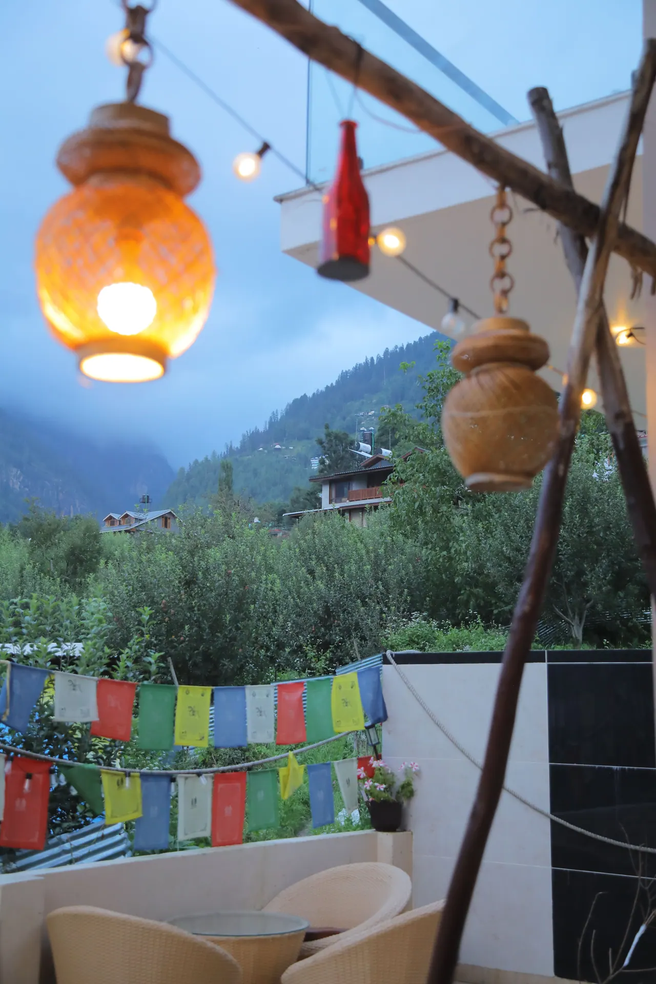 A glowing lantern and fairy lights over the terrace at dusk at Cafe DO NTHNG, with prayer flags and the Manali valley behind